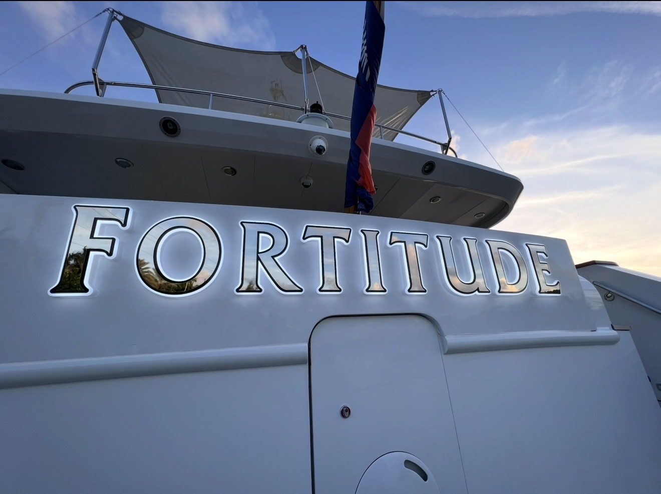 FORTITUDE in mirror-polished stainless serif letters with a white halo glow on a white yacht transom at dusk