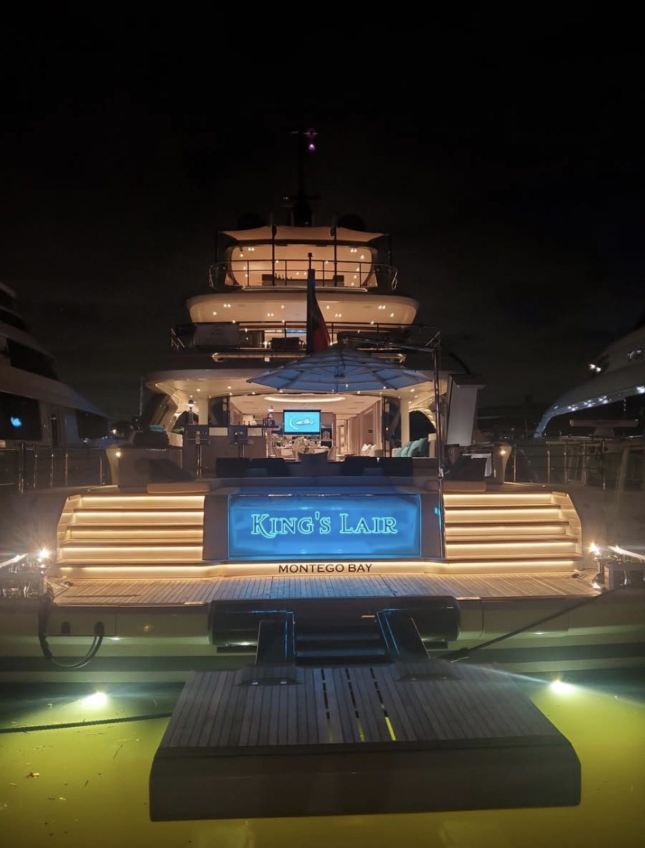 KING'S LAIR with MONTEGO BAY home port in teal-lit letters on an aft transom panel at night, with lit steps and underwater lights