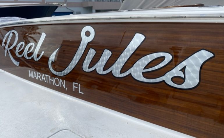 Reel Jules with MARATHON, FL home port in silver script letters on a varnished wood-grain hull side, daylight